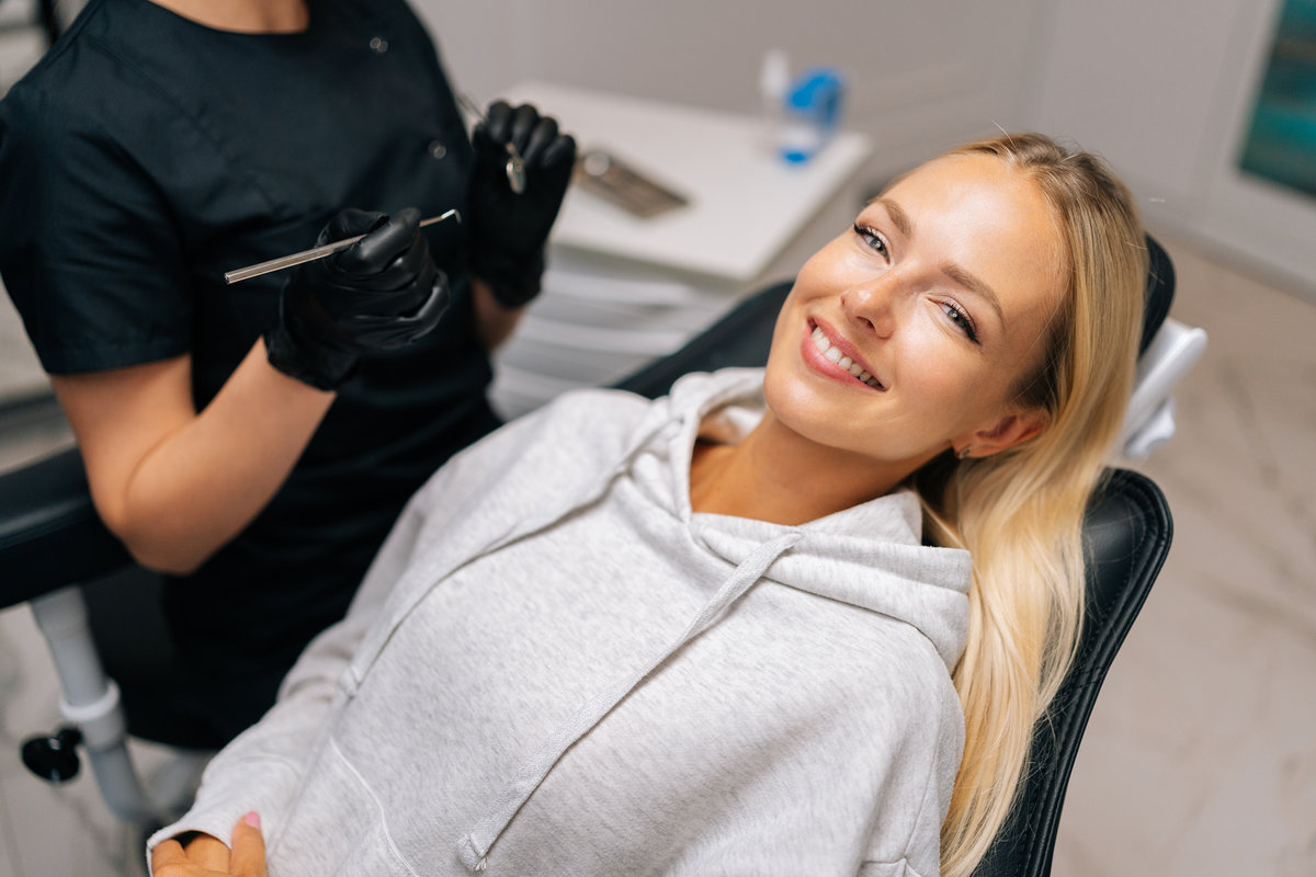 Portrait of charming young woman patient with perfect white teeth and smile satisfied after dental treatment lying on dental chair in dentistry clinic. Female dentist in gloves examining teeth.