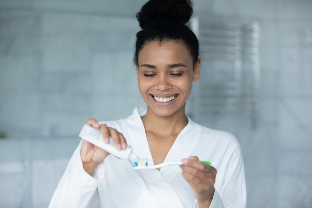 Happy young African American woman squeezing mint toothpaste from tube on plastic toothbrush for brushing teeth in bathroom, keeping morning routine for dental care, healthy enamel, mouth hygiene